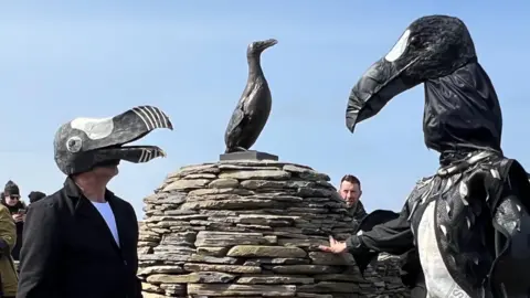 Two men dressed in black and white wearing headpieces modelled on the Great Auk stand next to an egg-shaped dry-stone cairn with a bronze statue of a Great Auk placed on top of it. Other people stand nearby.   