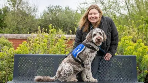 Batsonelli Photography Anne Herbert is standing behind a park bench. Her support dog Rafa is sitting in front of her on the bench. He is a labradoodle with grey and black markings.