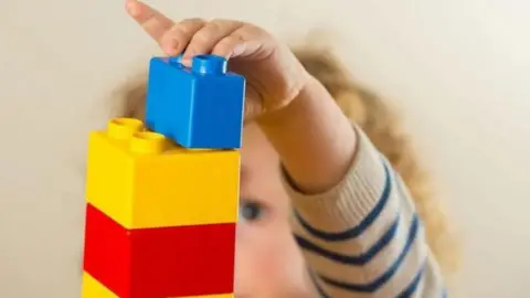 A girl with curly blonde hair and wearing a cream and blue striped jumper places a blue Duplo block on top of a yellow Duplo block on top of a red Duplo block. 