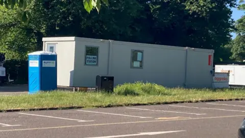 A portable cabin with 'polling station' posters in its window, and a portable toilet outside