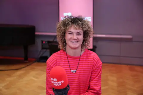  Ellie Kildunne smiles at the camera. She is wearing a red striped, long-sleeve top. She sits in-front of a red BBC Woman's Hour microphone.