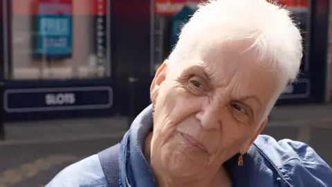 Linda Beddow is an older lady with short white hair wearing a blue coat and gold dangly earrings with red and blue stones. 