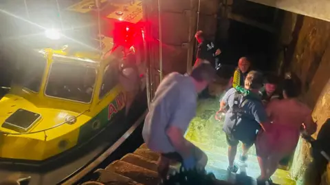Jake Magell A yellow-and-green rescue boat with bright lights and flashing red emergency lights is docked beside a stone stairway at night. Several people are moving down the wet steps from a waterside entrance while emergency responders stand near the boat and the opening in the stone wall. The scene is illuminated by intense rescue lights, reflecting off the water and highlighting the narrow harbor access point. Visible text on the boat includes “RESCUE” and partial lettering “AM.”