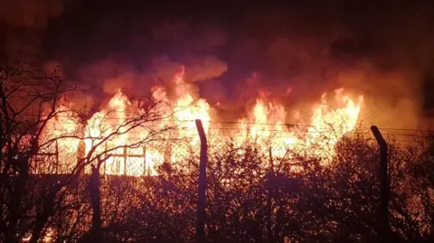 Liam Eli A large fire can be seen coming from a building at night time. A fence can be seen in front of the blaze and smoke billows into the night sky.