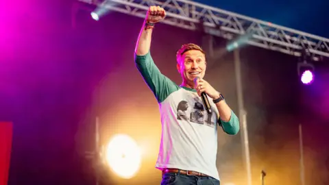 Ceremonia de victoria/Isla fuerte Russell Howard levanta su puño en señal de victoria en el escenario durante su presentación. Lleva una blusa de manga larga y jeans.