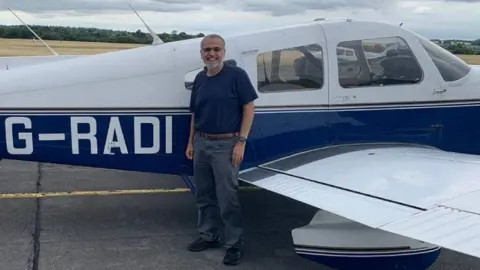 A man with a beard and sunglasses is seen standing in front of a blue and white plane