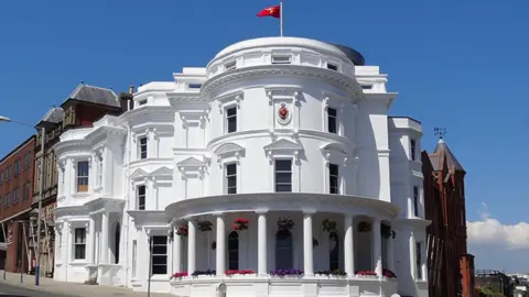 Manx Scenes The white parliamentary building has a semi-circular shape on the roadside corner, with pillars on the ground floor and a Manx flag on the roof. Due to its shape it is known as the wedding cake building.