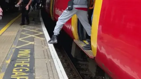 A photo of a man stepping off the train carriage, one leg is in mid air and the other on the train carriage, again there is a Mind the Gap signage on the platform floor.