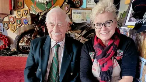 Julie Cooper who has blonde, curly hair, black glasses and is wearing a red, flowery scarf. She's sitting next to a man with white hair and a white moustache. He's wearing a blazer and green striped tie. Various plaques can be seen on the wall behind.