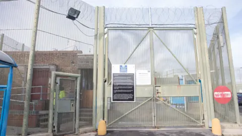 A prison gate with barbed wire and fencing running alongside. Various signs saying 'no entry' and 'HMP Erlestoke' also evident.