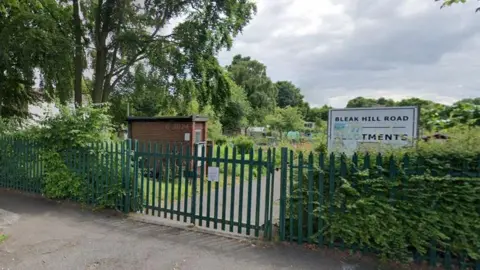 A set of allotments behind a green metal fence. Small huts, greenhouses and garden plots can be seen behind the fence. A white sign says "Bleak Hill Allotments".