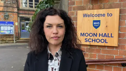 Michelle Catterson, Executive Head at Moon Hall School in Reigate, stood in front of a wall featuring a sign welcome people to the school. Michelle has long black hair and is wearing a black suit jacket and white patterned shirt. 