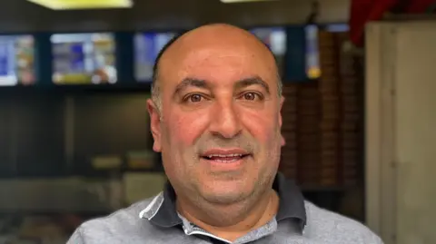 A head and shoulders image of Nuri Ozmicco. He is wearing a grey polo shirt and smiling while looking into the camera.
