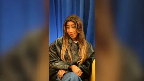A woman with brown hair and a black jacket sits in a chair.