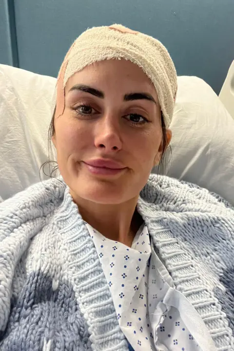 Lauren Macpherson Lauren Macpherson in a hospital bed. She has a bandage around her head, brown eyes and brown eyebrows. She wears a blue knitted cardigan over a hospital gown and smiles at the camera. 