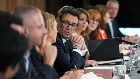 Christopher Furlong / Pool via AFP Andy Burnham clasps his hands as he looks towards camera while speaking to others sitting on a long table at a meeting of the National Economic Council in Manchester on 24 July.