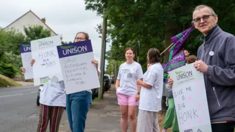 Striking NHS staff hold placards and flags on a residential road. One reads "honk for the NHS" and another says "save Aldeburgh Hospital kitchen and cleaning teams". The group are all women other than one man. It is a cloudy day and there are trees and a house next to the road.