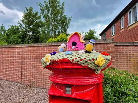 Shaun / BBC Weather Watchers Woollen figures including a couple of birds and a bird box, knitted from wood, adorn the top of a red postbox which is in front of a brick wall.
