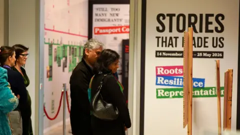 People walk through an art gallery. Stories That Made Us – Roots, Resilience, Representation, is written on the wall.