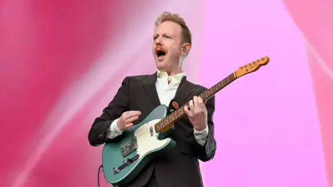 Two Door Cinema Club performing at Reading Festival, with singer/guitarist Alex Trimble shouting out. He is dressed in a suit and holding a guitar.