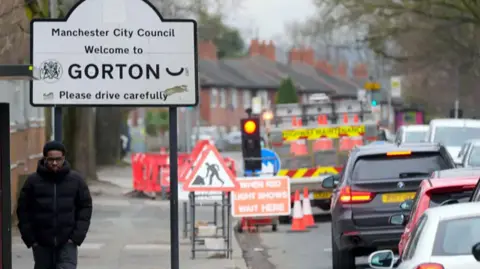 PA Media Generic street shot featuring a sign denoting the district of Gorton. There is a traffic jam and a teenager wearing a black puffa jacket, jeans and trainers walking along the pavement.