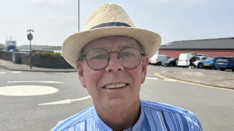 An older man wearing glasses and a straw hat. He is smiling at the camera and wearing a blue stripey shirt. He is standing beside a road in an urban area with the sea in the distance.