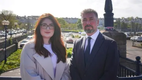 Alliance A man and a woman stand side-by-side near the Queen's Parade area of Bangor.
The woman has long reddish hair and is wearing glasses, a light coloured white T-shirt and overcoat. The man next to her is wearing a dark suit and blue tie. He has light stubble and short hair.