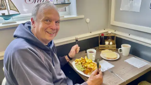 A man in a navy hoodie sits at a cafe table with a plate of fish, chips, mushy peas, gravy and a pie. There is also a side plate of butter and a cup of black tea. There is a window beside him with two toy boats resting on the sill.