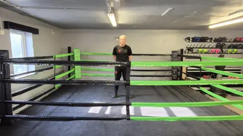 Damon Croft A man standing a boxing ring. He is wearing a black T-shirt and trousers and has his hands behind his back. The boxing ring is green and black.