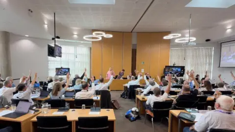 Zoie O'Brien/BBC A meeting room at West Suffolk Council's HQ. There are rows of tables with people sitting at them, facing away from the camera. They are all raising their arms in a vote. At the far end of the room, there are at least five people at a desk, facing into towards the camera.