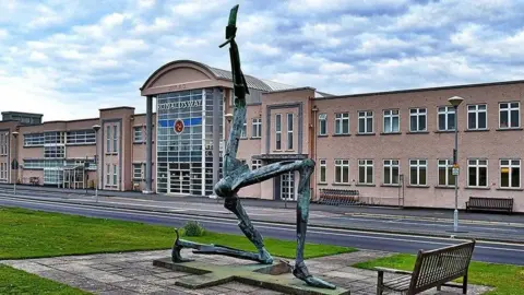 A three legs of man statue outside the entrance of Ronaldsway, which is a peach coloured block building with a rounded roof in the centre and a glass-fronted entrance with Ronaldsway on it in white lettering.