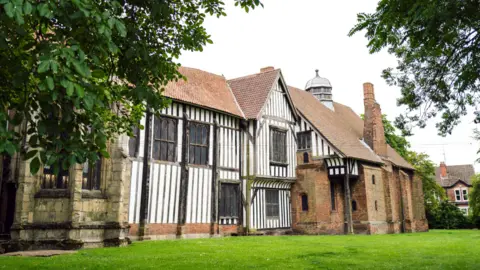 English Heritage An outside view of Gainsborough Old Hall. It has a white and black exterior as well as orange and beige brick. Trees cascade over the top of the picture.