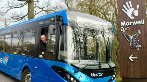 Brown sign for zoo on right with monkey logo and blue bus to the left with driver at the window.