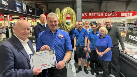 BBC Kevin Parsons being handed his long-service certificate. He is stood in the freezer section of the supermarket, surrounded by other members of staff