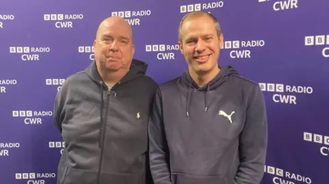 BBC Neil Jeffs on the left is standing next to his younger brother Paul, who smiles at the camera. They are opposite the BBC Radio CWR brand. Neil is bald and wears a light blue hoodie. His brother has short, dark to gray hair, smiles at the camera, and is also wearing a light blue hoodie.