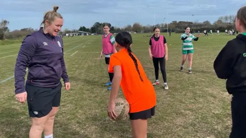 BBC England women's rugby captain Marlie Packer talks to a girl wearing an orange t-shirt as the girl prepares to pass a rugby ball to another player. Marlie is wearing an England training jumper and shorts.