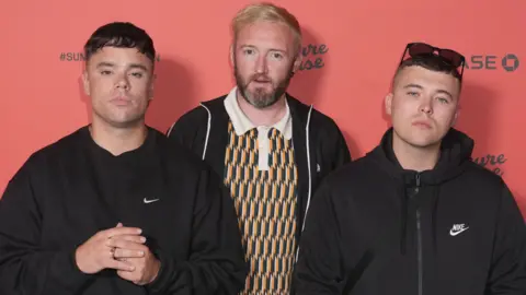 PA The three band members looking at the camera. One has sunglasses on his head with a dark hoodie. One is wearing a ring and a dark jumper. The other member stnds behind the first two and is wearing a colourful patterned top