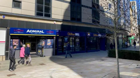 People walking past the entrance to a 24-hour slot machine arcade on a street with a couple of trees and more betting shops further down the same parade
