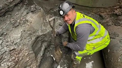 S.I UXO A man in a high-vis jacked crouches in a muddy pit. He is holding a shovel in one hand. Next to the shovel is the WWII bomb in the mud.