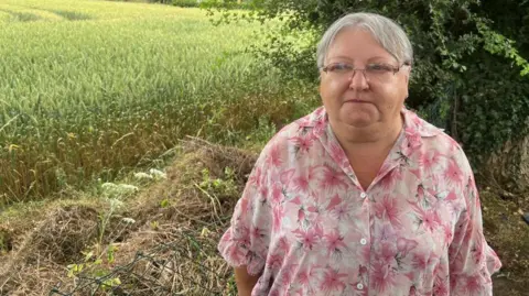 The photograph shows a woman standing at the edge of a field beside a line of trees and hedgerow. She is wearing glasses and a loose-fitting pink floral shirt.
A large field of cereal crops stretches across most of the background.