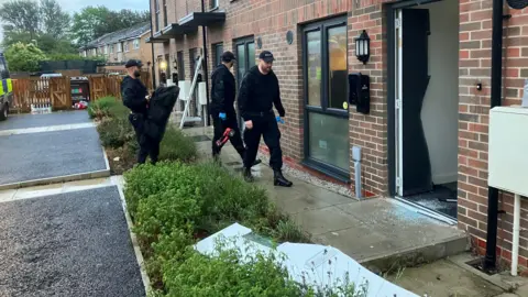 Photograph of the aftermath of drugs raids on Hopwood Street in Pendlebury. the image shows police officers and a door which has been removed from its hinges. 