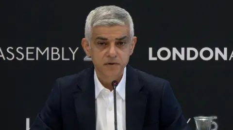 Sadiq Khan is seen speaking at mayor's question time with a mic in front of him. London Assembly branding is visible behind him.