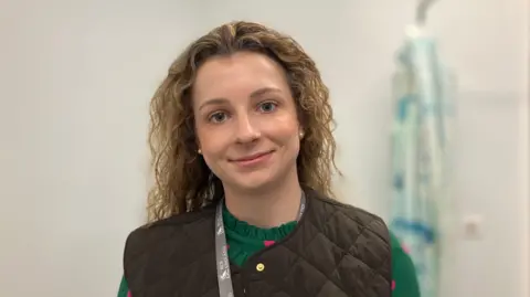 Olivia Howe looking at the camera, standing in a pale coloured room with a hospital gown hanging behind her