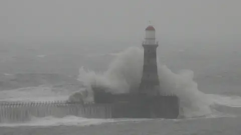 BBC Weather Watchers / Gill Helps A white-and-red lighthouse at the end of the pier can just be made out through the mist. A white, frothy wave crashing into the pier rises about halfway up the structure.