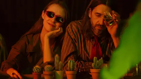 Factual Fiction A woman wearing sunglasses sits next to a man holding a glass. They are sitting behind 5 cacti in plant pots on a table.