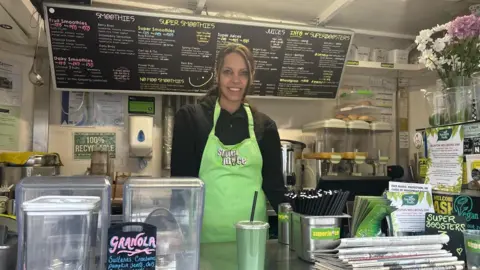 BBC Alex Frost, the owner of Super Juice, standing in her van.  She is wearing a black top and a green apron with Super Juice written on it.  She is standing in front of a menu board.