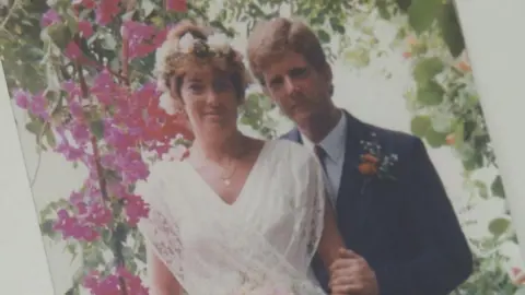 A photo of Joyce and Mike Gifford in wedding attire. She is wearing a white, sleeveless lace dress with a white flower crown.He is standing behind her with his hand on her arm and is wearing a dark suit and tie.