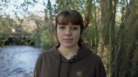 Molly Hadley wears a brown hoodie, grey t-shirt and has her  dark brown hair tied up in a ponytail