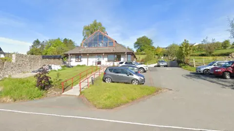 Google Maps A white GP surgery with a brown tiled roof and glass window. There are concrete steps leading up to the front door, with red railings. Surrounding the step is green grass. To the right of the surgery is a car park with parked cars. In the background of the picture are green trees and a field.