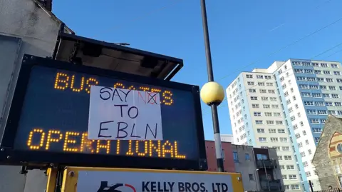 A paper notice reading "say no to EBLN" has been pasted over an LED notice board which reads "bus gates now operational".